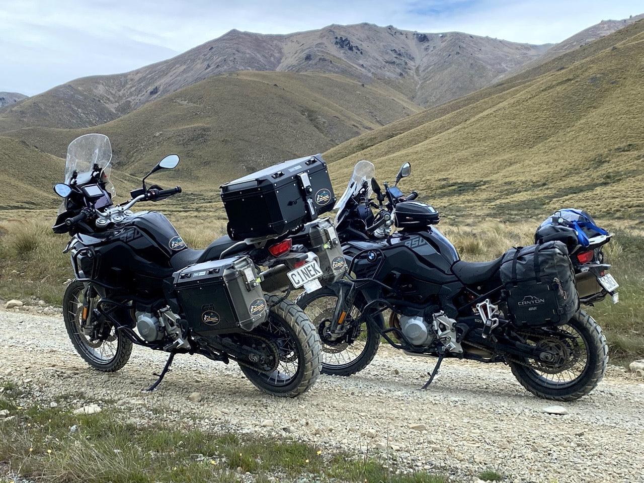 Two BMW motorcycles with luggage parked on a gravel road against a mountainous backdrop